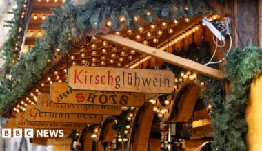 Wooden signs lined up over a wooden stall, in German , advertising wares such as Kirschgluhwein, shots and German beer. The stalls are adorned with fir tree garlands and have light garlands strung up.