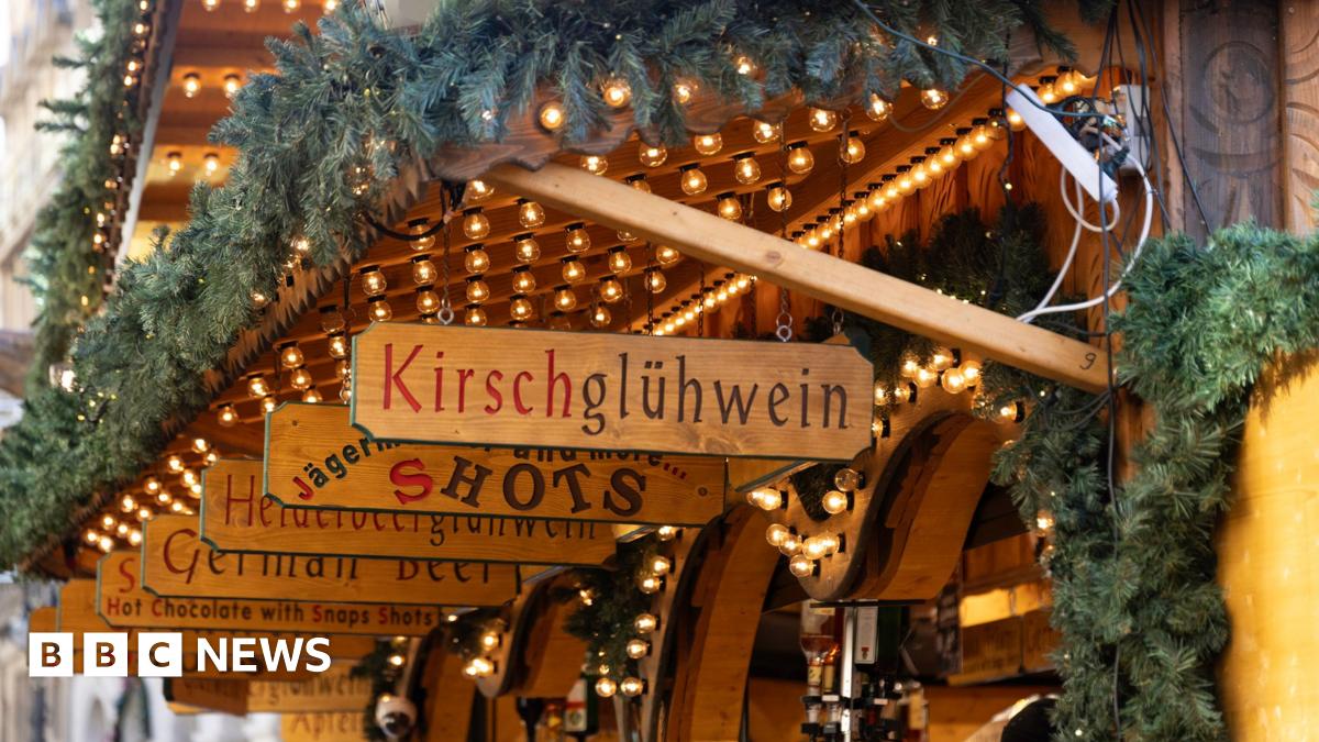 Wooden signs lined up over a wooden stall, in German , advertising wares such as Kirschgluhwein, shots and German beer. The stalls are adorned with fir tree garlands and have light garlands strung up.
