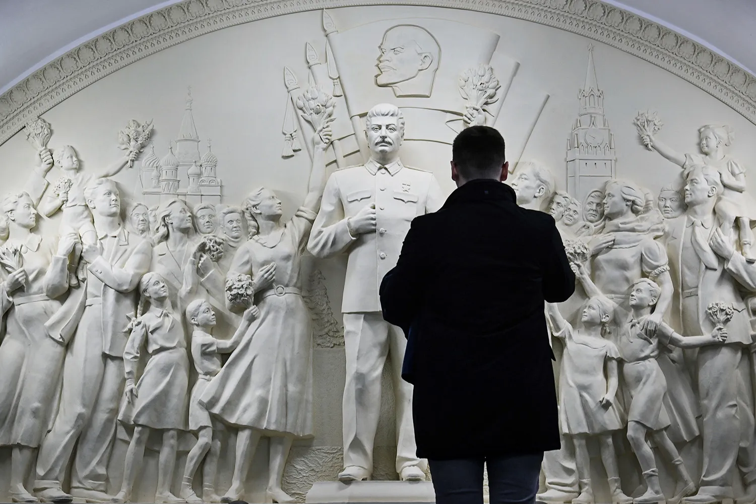 A person is silhouetted in front of a white high relief statue of Stalin with other figures around him.