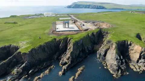 PA Media An aerial image of SaxaVord spaceport under construction on the island of Unst in Shetland. There is a launch pad with a launch tower on a flat area of land above sheer cliffs to the sea below.