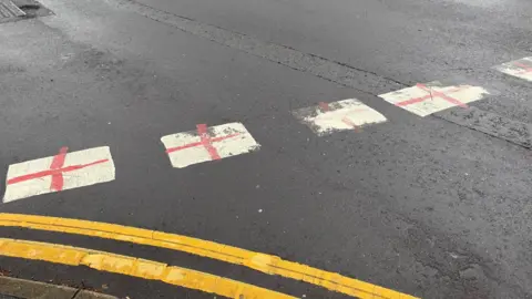 BBC/Louis Reynolds A close-up of a grey tarmac road, with bright double yellow lines in the foreground. In the centre of the frame are white give way road markings, which have been covered with red crosses.