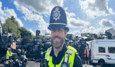 Sgt David Eeles pictured smiling while stood in a scrap yard. He is wearing a black police helmet and police yellow jacket