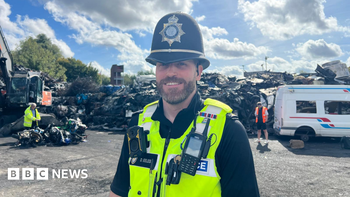 Sgt David Eeles pictured smiling while stood in a scrap yard. He is wearing a black police helmet and police yellow jacket