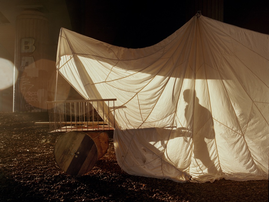 Photo of a figure pushing a cart; the figure is visible in silhouette behind a large white sheet.