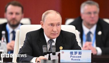 Vladimir Putin, wearing a dark suit and black and white tie, sits at a desk at a summit speaking into a microphone. A sign in front of him at the desk states the word "Russia" in Chinese, Russian and English.
