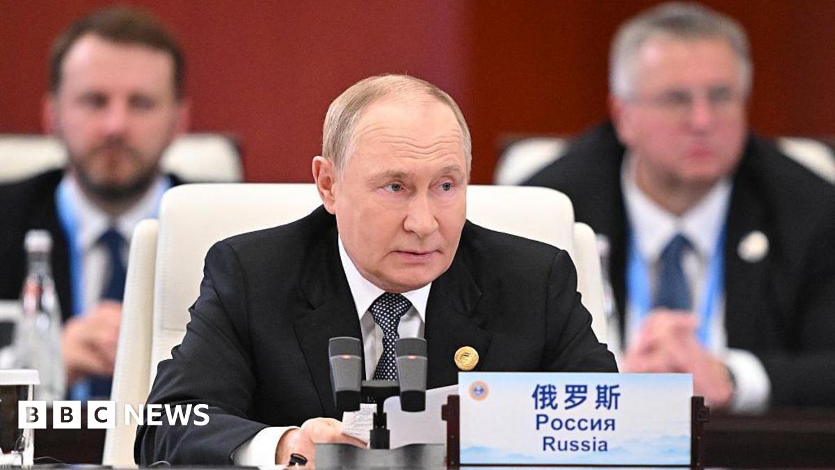 Vladimir Putin, wearing a dark suit and black and white tie, sits at a desk at a summit speaking into a microphone. A sign in front of him at the desk states the word "Russia" in Chinese, Russian and English.