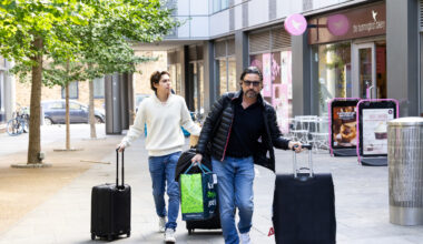 Two people wheeling large suitcases outside of a dorm building in London.