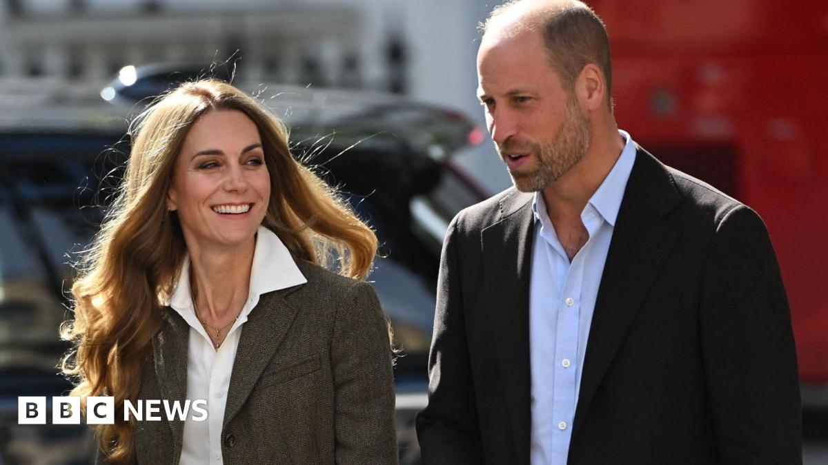 Shoulder crop of William and Catherine. William is wearing a black blazer and light blue shirt. Catherine is wearing a tweed-style jacket and white shirt. She has blonde hair. Both are smiling and posing outside the museum.
