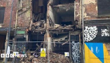 Metal fences sit at the feet of a collapsed building. Fallen brickwork and debris can be seen behind.