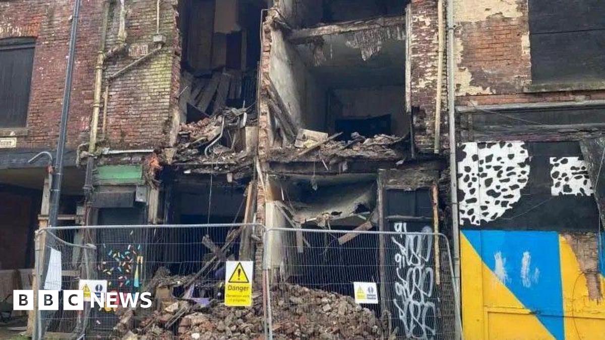 Metal fences sit at the feet of a collapsed building. Fallen brickwork and debris can be seen behind.