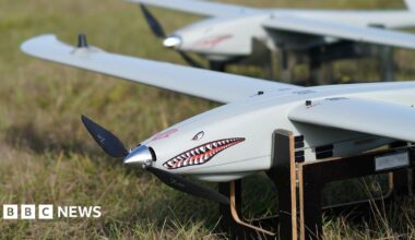 A Ukrspecsystems drone with a shark face and design on its note propped up on a wooden stand in a field.
