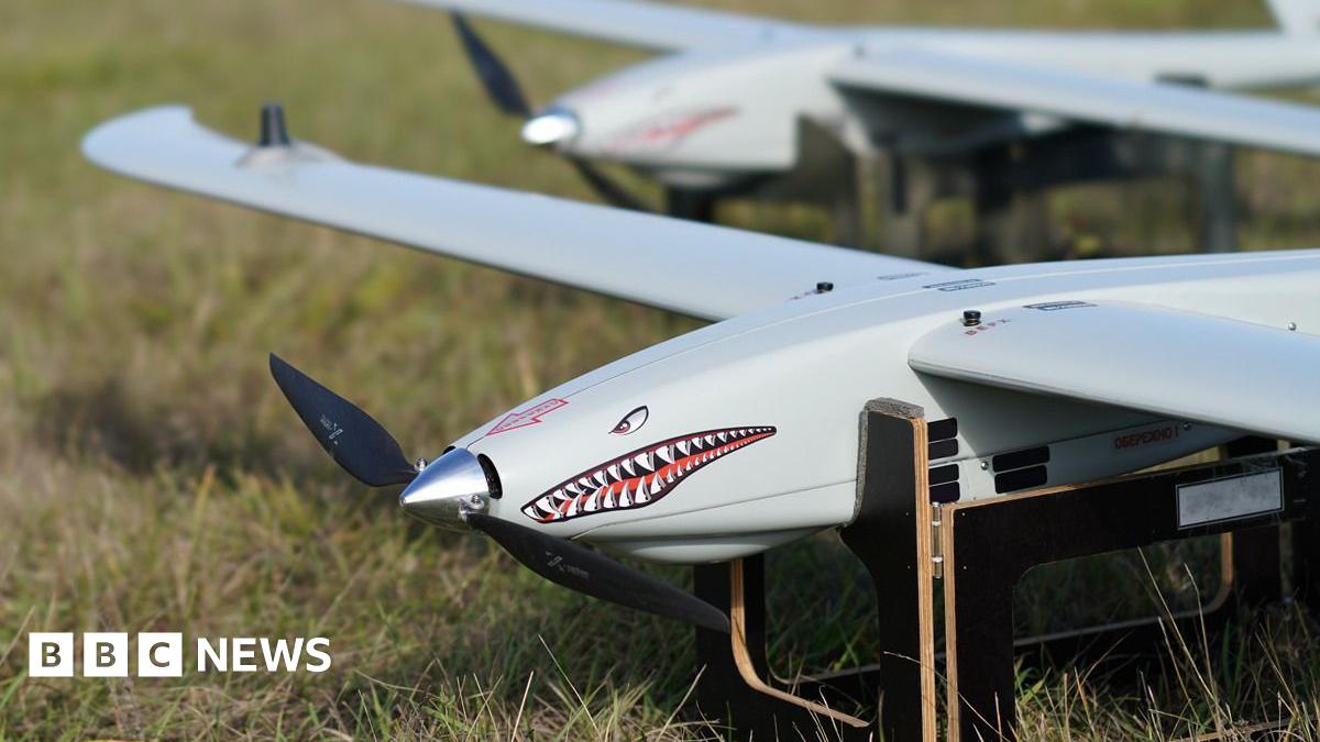 A Ukrspecsystems drone with a shark face and design on its note propped up on a wooden stand in a field.