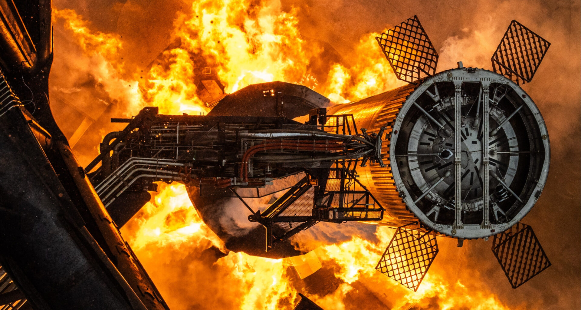 fire erupts from below a giant chrome cylinder with four grid fins, next to a tower.