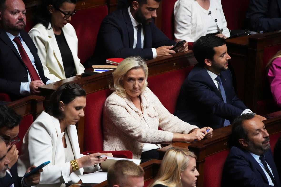 Several individuals sit in rows of red velvet benches, some reading papares on their desks, some listening to what is being said