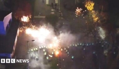 The image shows a stand off between police and young people on Hay Avenue in Niddrie on Bonfire night 2023. Police Scotland officers are standing in a line with police cars behind them. The police are blocking young people from entering the road. There are around 30 young people stood at the round about to the road.