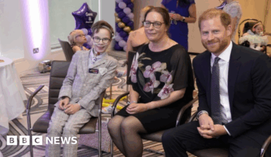 A boy in a grey suit sits next to a woman in a black floral dress, with Prince Harry sat next to them. The boy has a tube near his nose. They all smile at the camera.