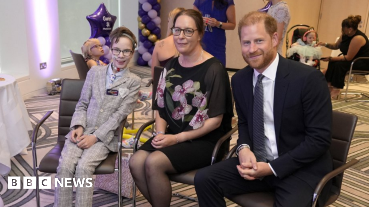 A boy in a grey suit sits next to a woman in a black floral dress, with Prince Harry sat next to them. The boy has a tube near his nose. They all smile at the camera.