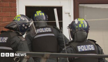 Three police officers enter a house. They are wearing helmets and other protective clothing.
