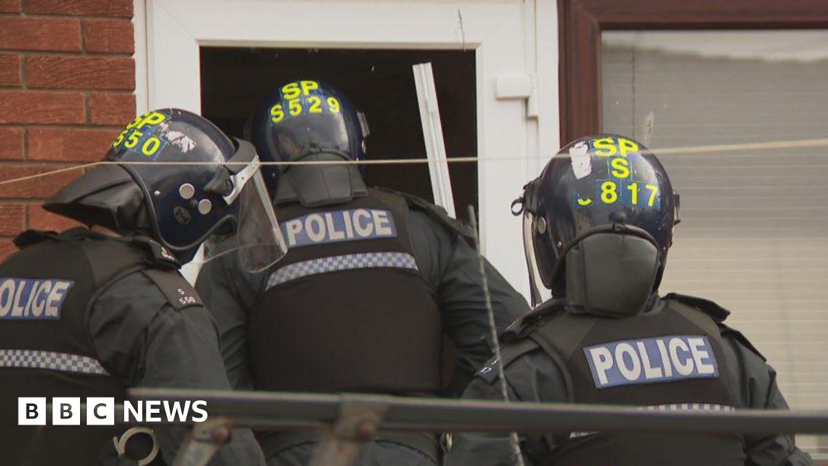 Three police officers enter a house. They are wearing helmets and other protective clothing.