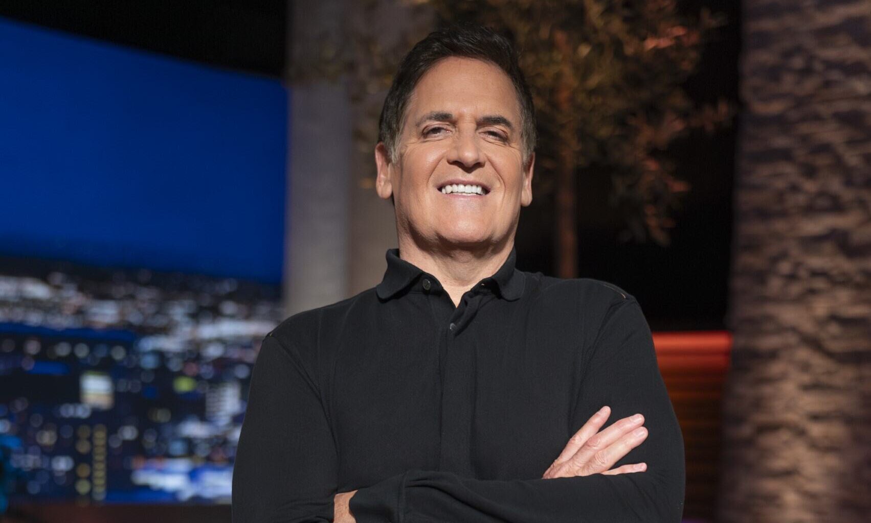Mark Cuban in a black shirt on set smiling with his arms crossed