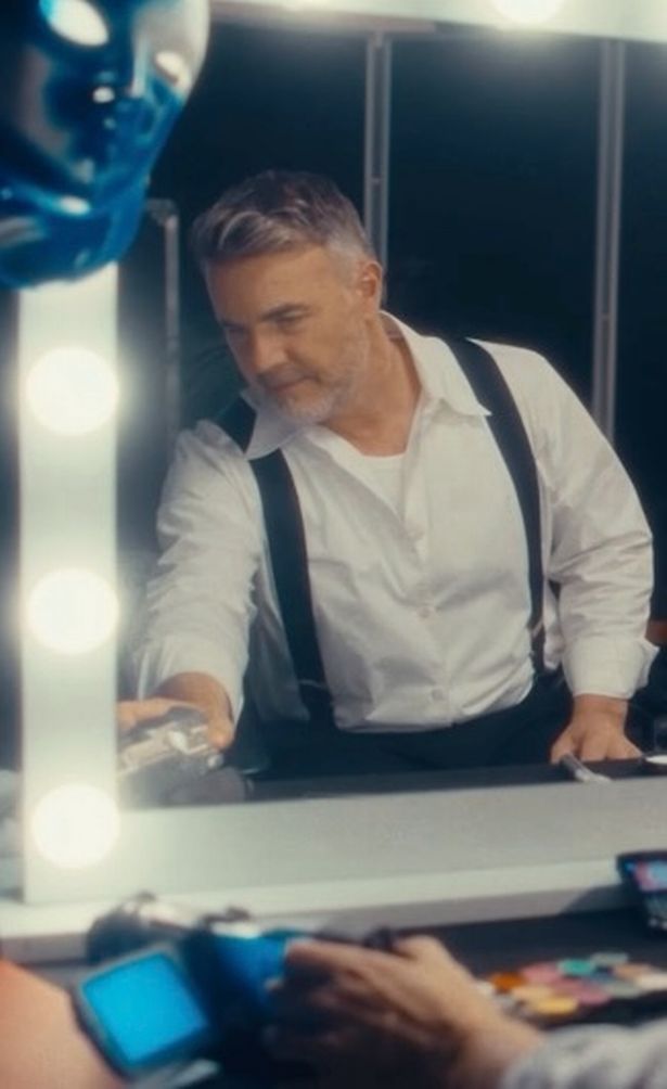 Gary Barlow in a white shirt and dark trousers in front of a mirror.