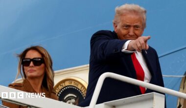 Donald Trump is wearing a navy suit jacket, white shirt and red tie. He is standing at the top of a staircase leading to the blue Air Force One jet. He is pointing towards someone on the ground. Behind him, Melania Trump wears a brown jacket and dark sunglasses.