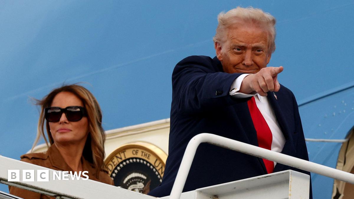 Donald Trump is wearing a navy suit jacket, white shirt and red tie. He is standing at the top of a staircase leading to the blue Air Force One jet. He is pointing towards someone on the ground. Behind him, Melania Trump wears a brown jacket and dark sunglasses.