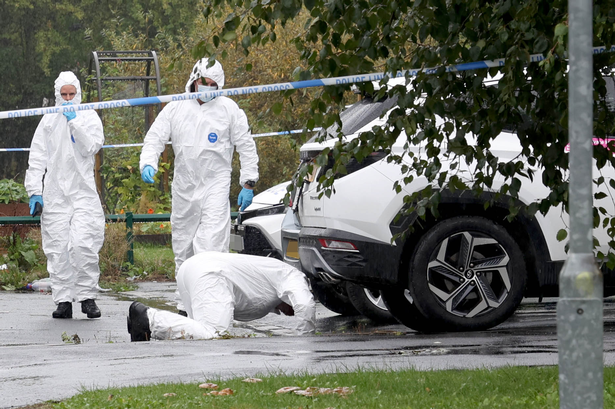 Forensics investigators search underneath cars following a shooting in Gorton