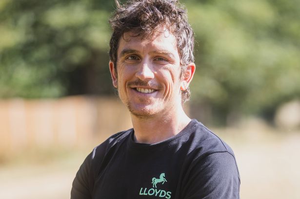 Thomas poses here with a black t-shirt. He's in front of a grassy backdrop and has his arms folded and is smiling at the camera slightly side on