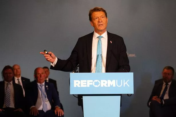 The image shows Mr Tice wearing a suit with a sky blue tie and addressing people from the stage at a press conference