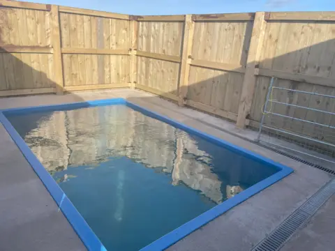 Caithness Seal Rehab and Release Centre A small rectangle-shaped outside pool made from blue plastic and sunk into the ground. The pool is surrounded by a concrete area and around that is a high fence made from wooden panels.