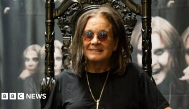 Ozzy Osbourne sits on a dark wood, high-backed chair with ornate designs on it. He is wearing round sunglasses, and is smiling, while also wearing a black T-shirt and a silver pendant chain. Behind him there are images of him on canvas with different facial portraits.