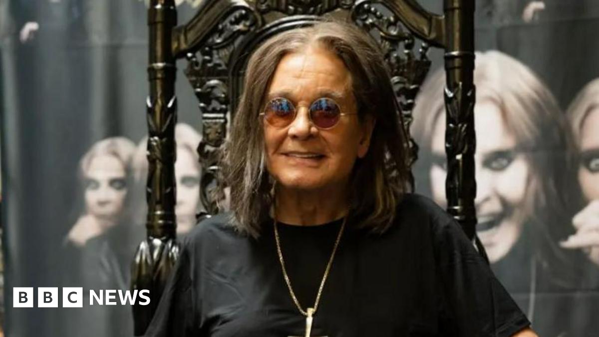 Ozzy Osbourne sits on a dark wood, high-backed chair with ornate designs on it. He is wearing round sunglasses, and is smiling, while also wearing a black T-shirt and a silver pendant chain. Behind him there are images of him on canvas with different facial portraits.