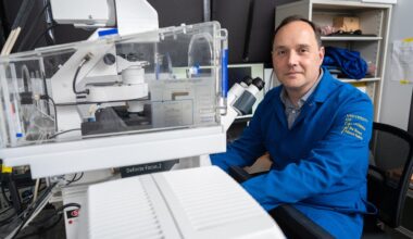 Researcher in a UC lab coat sits beside advanced microscope equipment, preparing for imaging work in the laboratory.