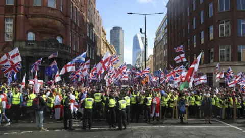 EPA A crowd of hundreds gathers on the junction of two London streets with tall City high rises visible in the background. There are tens of Union Jack and St George's flags. In front of the crowd stands a row of police officers in high vis jackets.