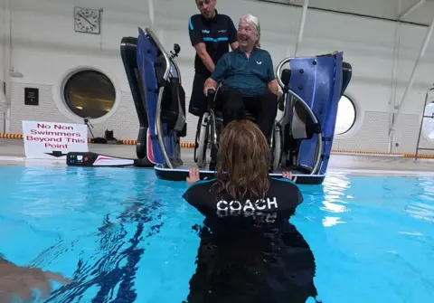 A view from behind the swimming coach as the PoolPod is lowered. We see Judith in the pod about to go into the water. There is a clock on the wall behind and a sign on the floor saying "No non swimmers beyond this point".
