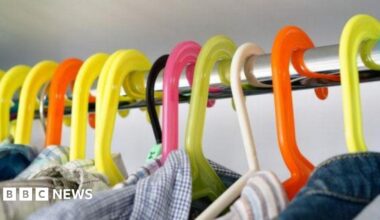 Different coloured clothes hangers on a metal pole. There are flannel tops on the hangers. The background is grey.