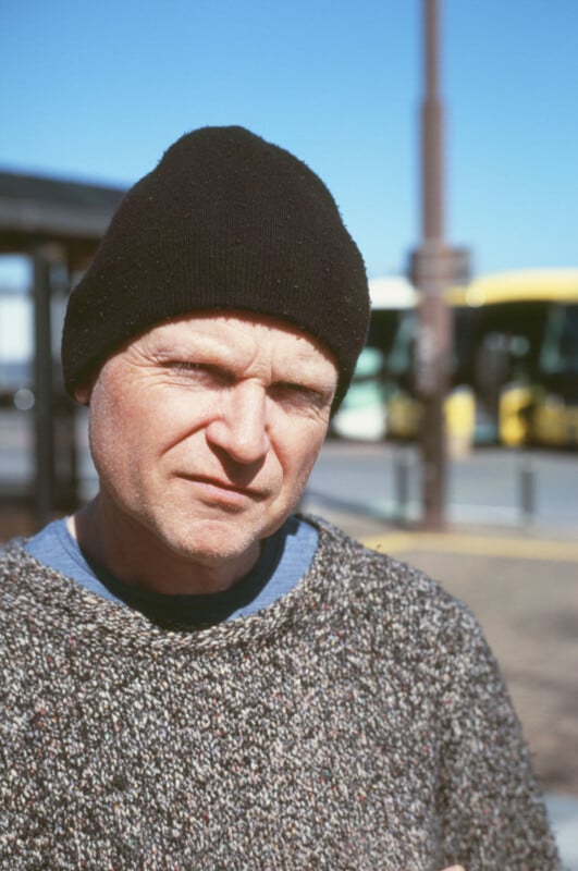 A man wearing a black knit beanie and a gray sweater stands outdoors on a sunny day, with blurred yellow buses and a shelter visible in the background.
