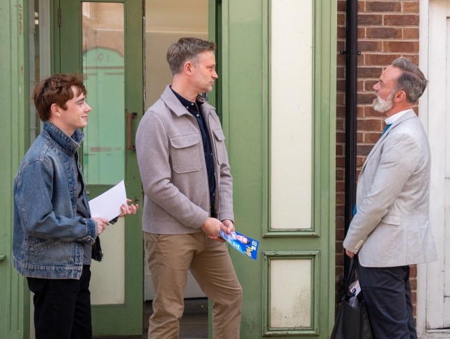 Billy talks to Noah and Miles, holding flyers, outside the community centre in Corrie