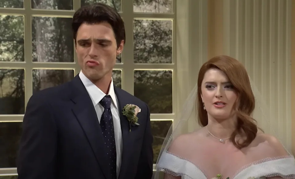 A couple in formal attire, possibly at a wedding. The man wears a dark suit with a boutonniere; the woman is in a bridal dress, looking slightly amused