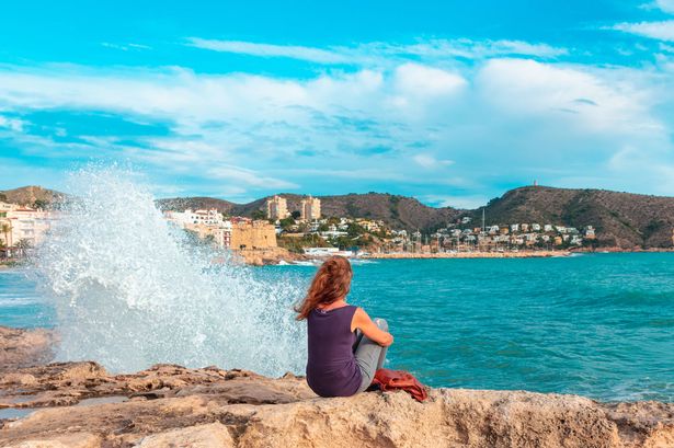 Woman sitting looking at waves crashing on rock ( Moraira,  Alicante province in Spain)
