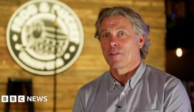 John Bishop is talking in this screenshot from the interview, with the blurred out Frog and Bucket logo and the stage and microphone seen behind him.