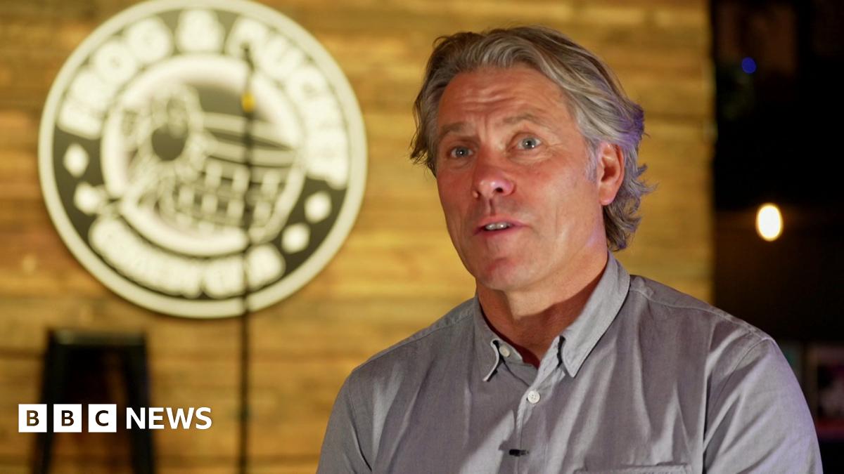 John Bishop is talking in this screenshot from the interview, with the blurred out Frog and Bucket logo and the stage and microphone seen behind him.