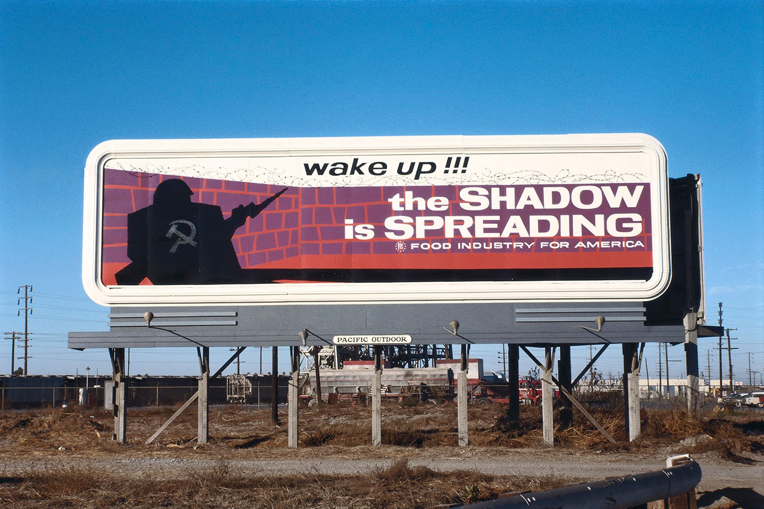 An anti-communist billboard with a hammer and cycle on a man’s silhouette with a gun in front of a brick wall reads "wake up!!! the SHADOW is SPREADING" with a logo for the food industry for America.
