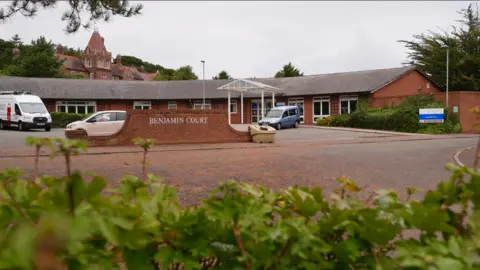 Martin Giles/BBC A wide view of Benjamin Court, showing the front car park fronted by a brick wall sign with Benjamin Court on it in white lettering. A single storey red brick building is beyond. One white transit van and two cars are in the car park.