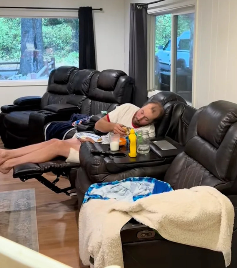 Person relaxing on a recliner in a cozy living room, holding a sandwich with snacks and drinks nearby