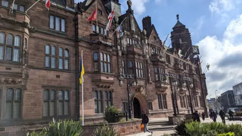 Ellie Brown Coventry City Council offices are in a historic building that was constructed in the 20th Century in an early Tudor style. The building has pedestrian areas outside and flower beds and there are flags decorating the building. People are walking around the grounds.
