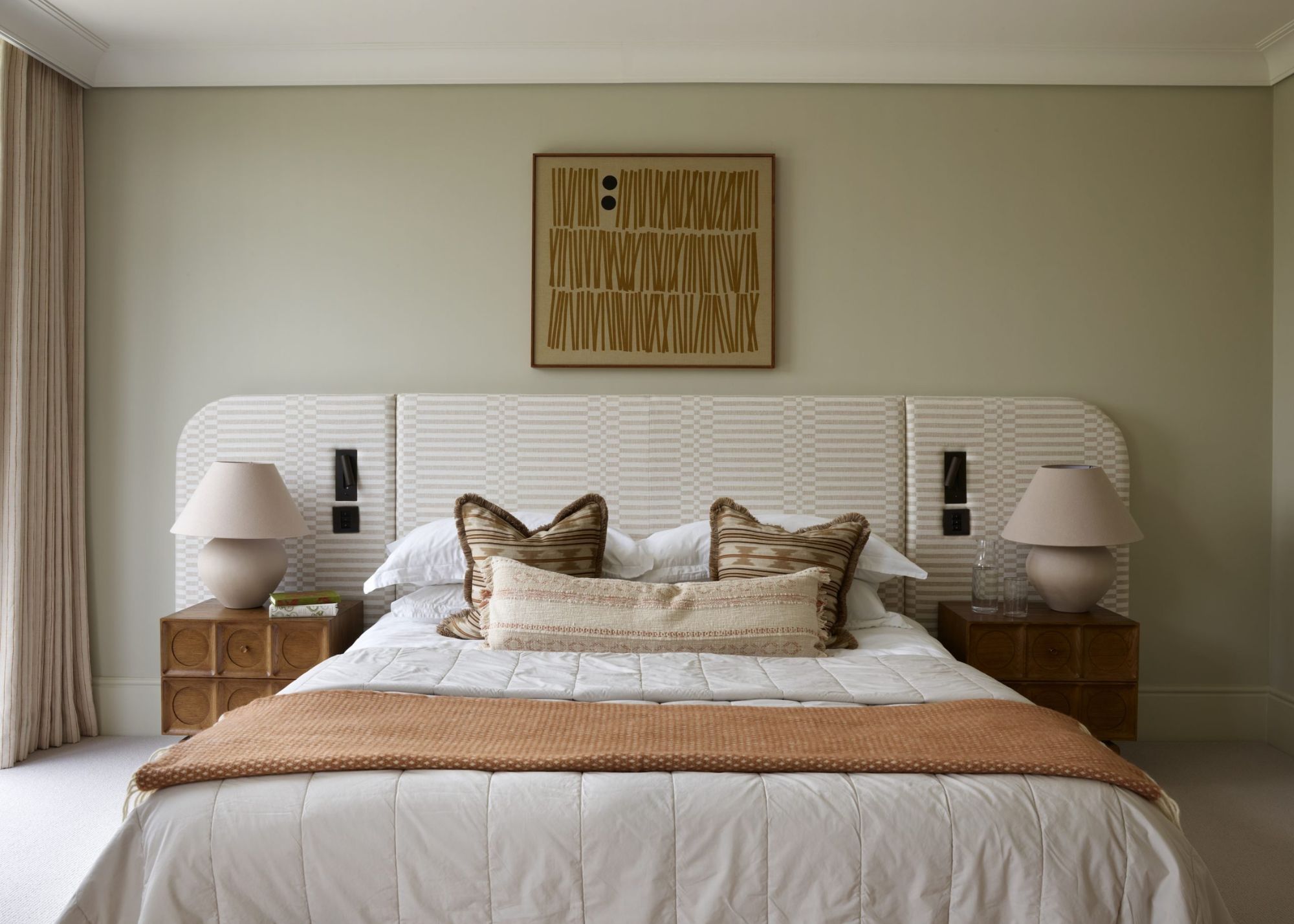 A modern bedroom with sage green walls, an elongated headboard, and neutral bedding.