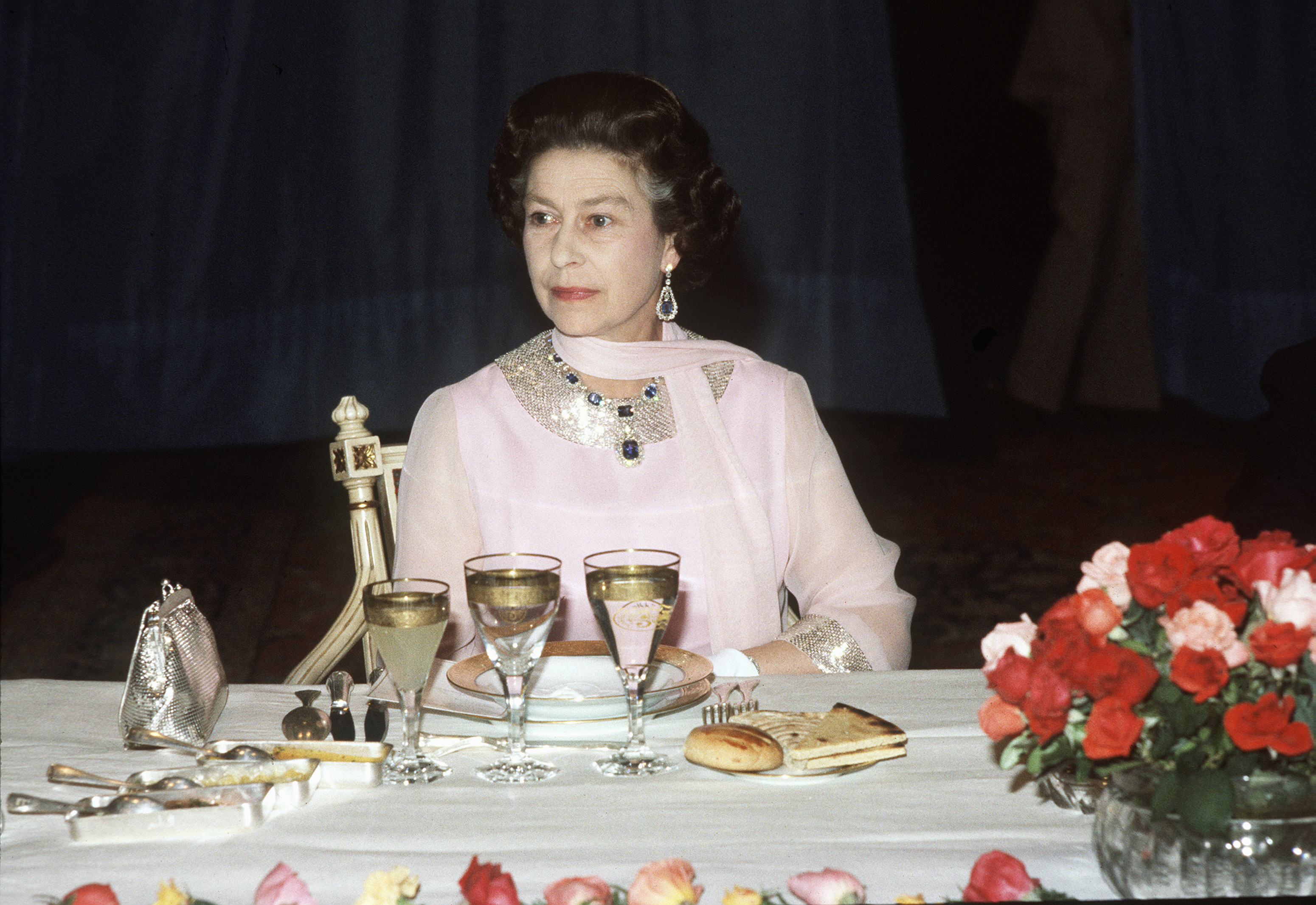 Queen Elizabeth wearing a pink dress and scarf sitting at a table with food and glasses