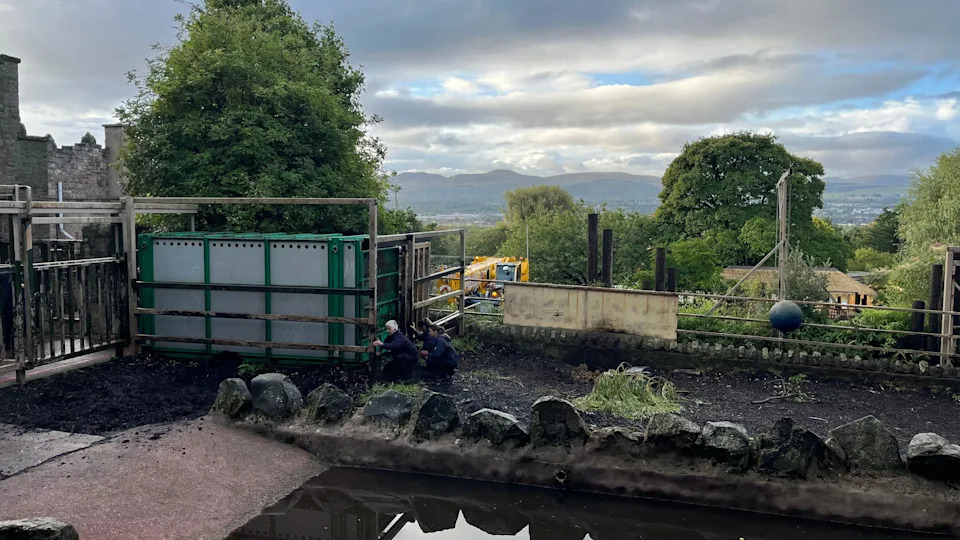 An empty animal enclosure at Edinburgh Zoo. Dark soil is on the ground. The sky is blue and grey with large streaks of grey cloud.